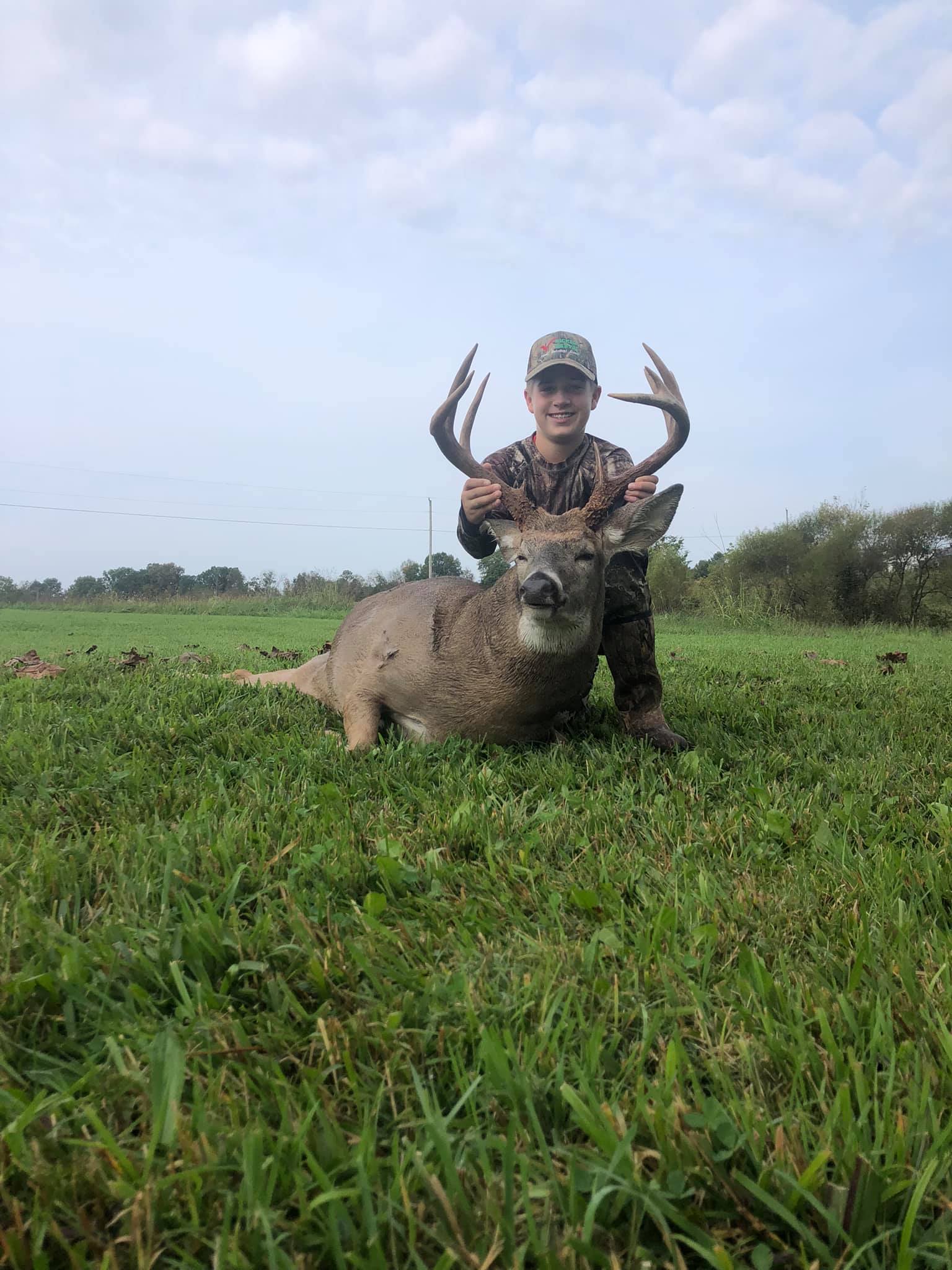 Young hunter poses with his large whitetail buck on a grassy field.