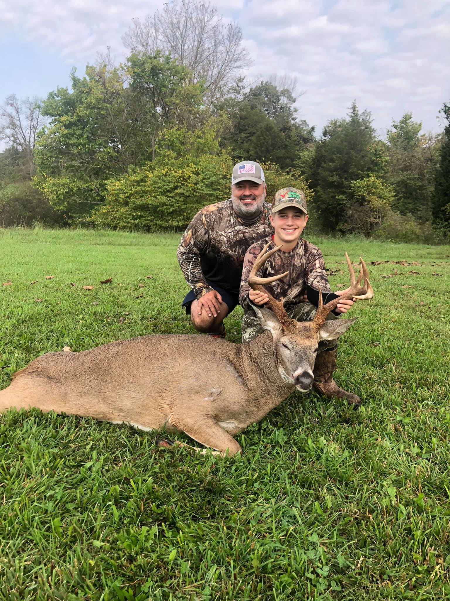 A smiling father and son with a harvested buck in a grassy field.
