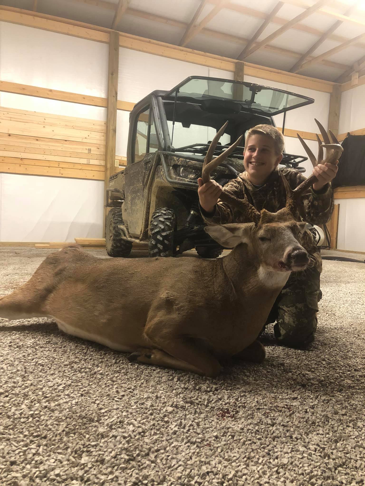 A young hunter kneels proudly with a harvested buck, showing off its antlers.