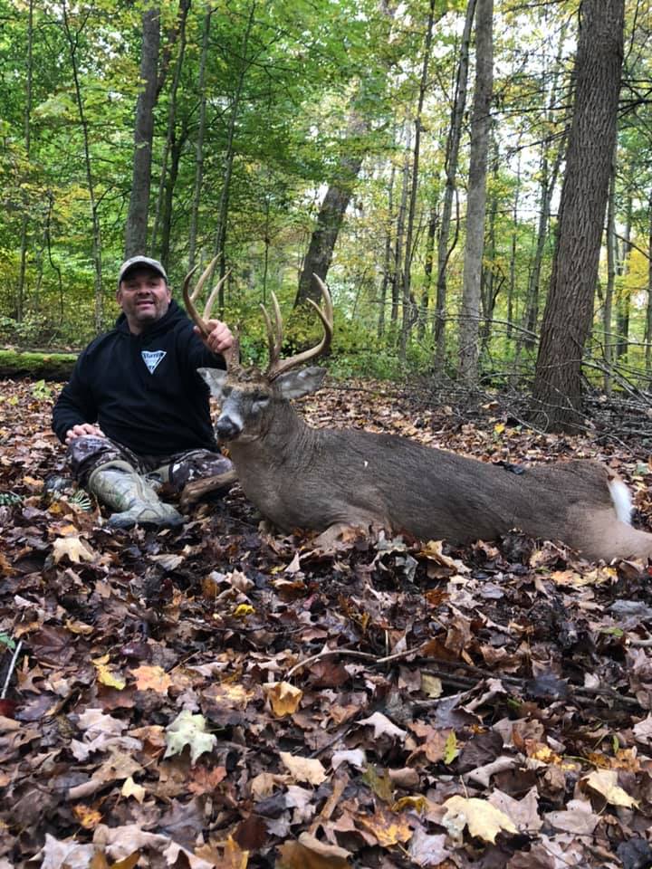 Man poses with a large buck deer in the woods during autumn.