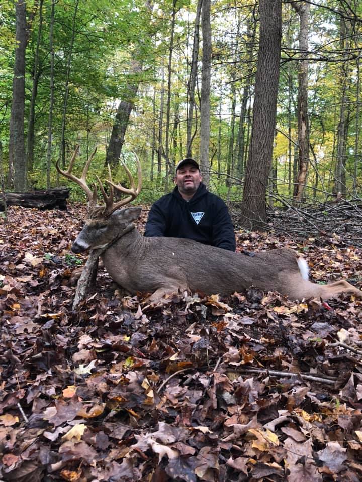 A hunter poses behind a harvested buck in a leaf-covered forest.