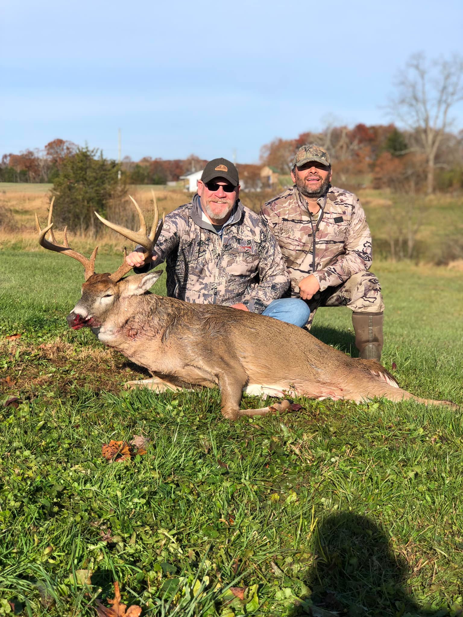 Two hunters pose with a buck on a grassy field with trees in the background.