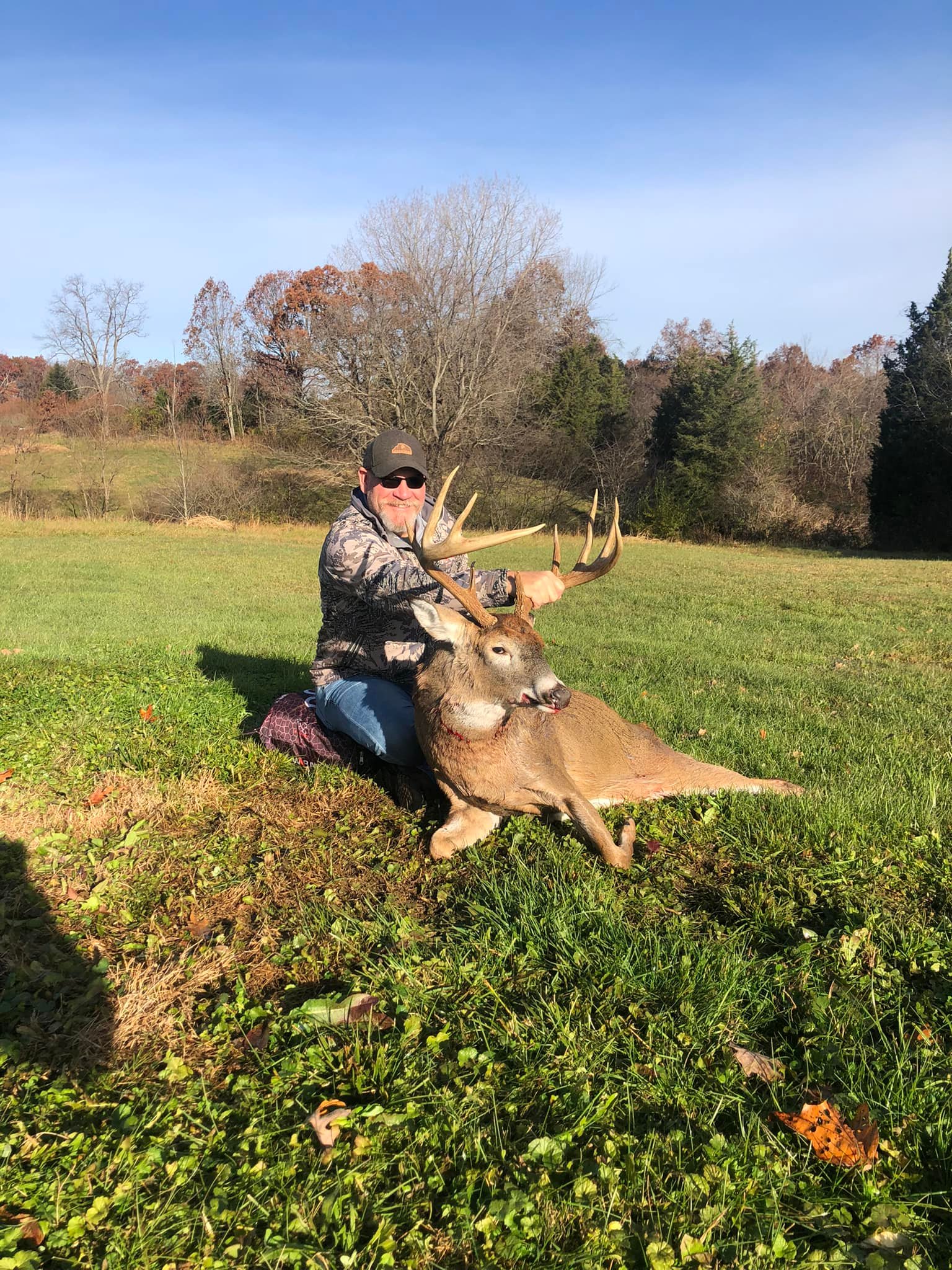 Hunter poses with a large buck in a grassy field with trees in the background.