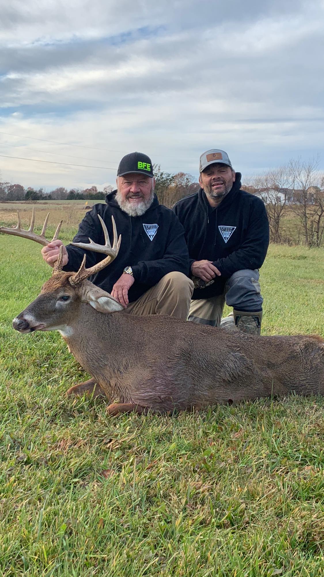 Two hunters pose with a mature whitetail buck in a grassy field.