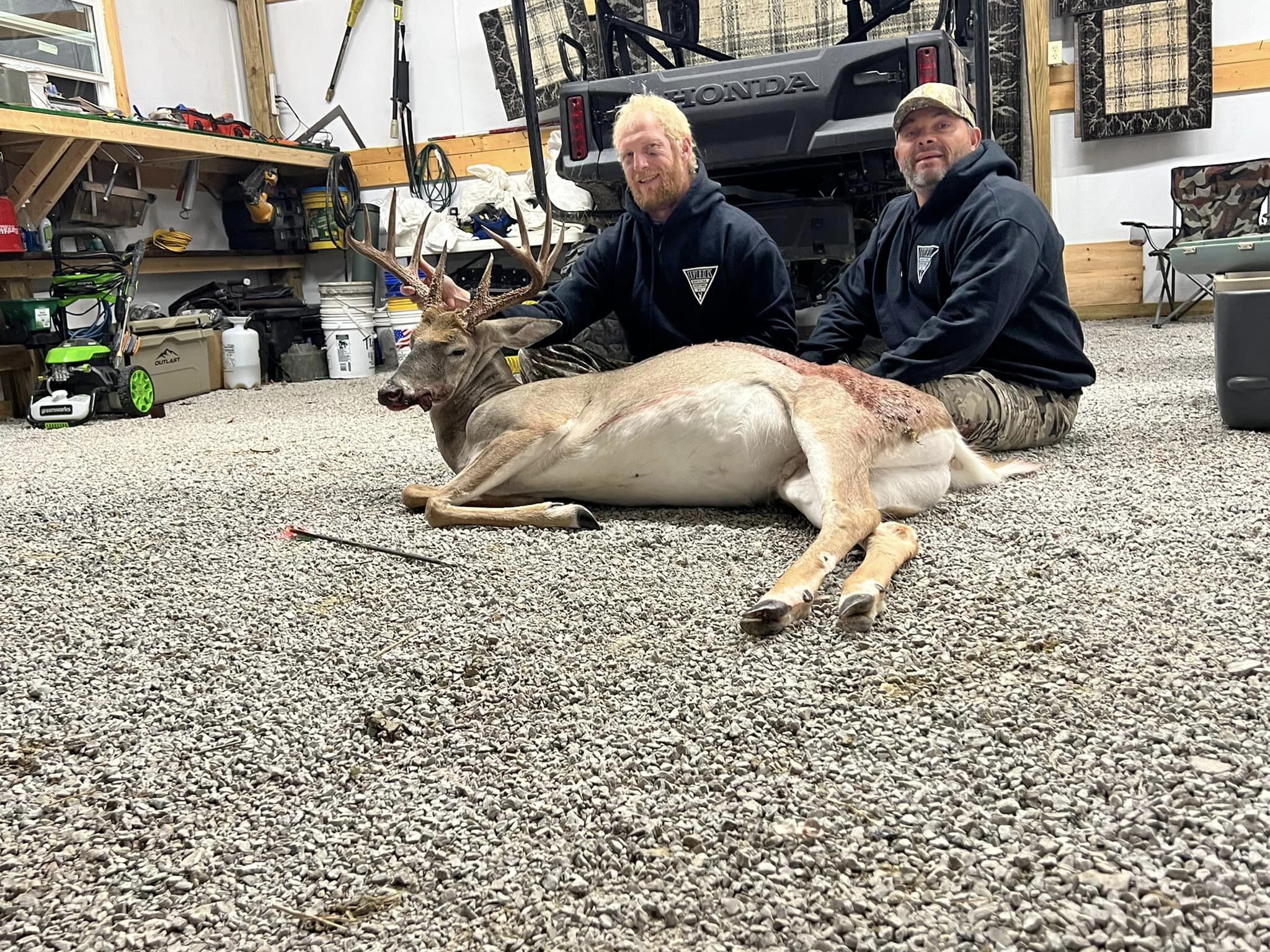Two hunters with a harvested buck in a gravel lodge with a Honda ATV.