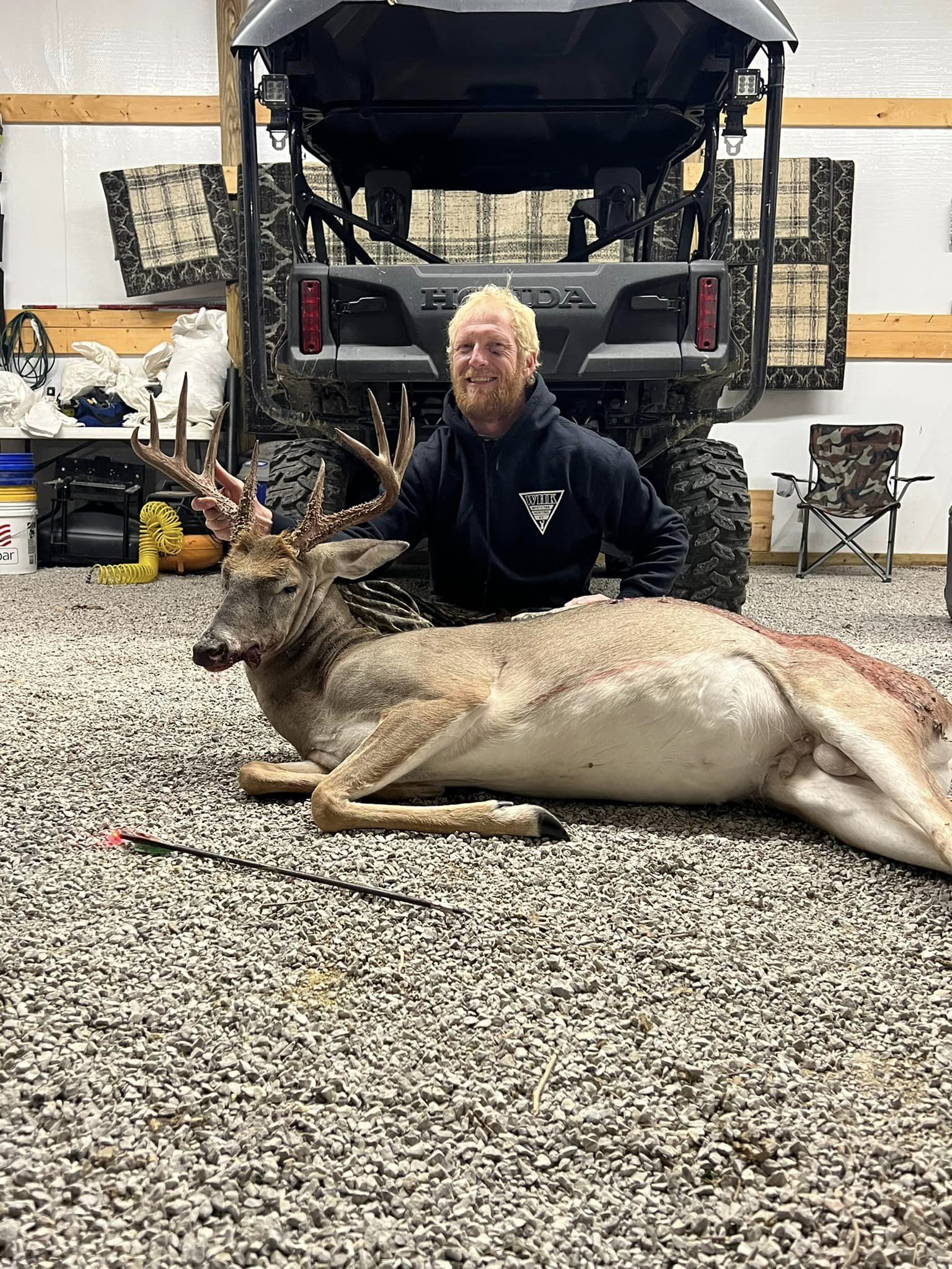 A hunter smiles behind a mature buck he harvested, with an arrow in the ground nearby.