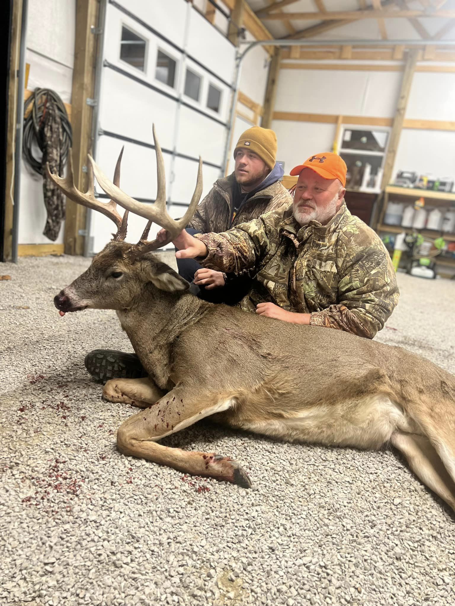 Two hunters proudly display a large buck in front of a garage.