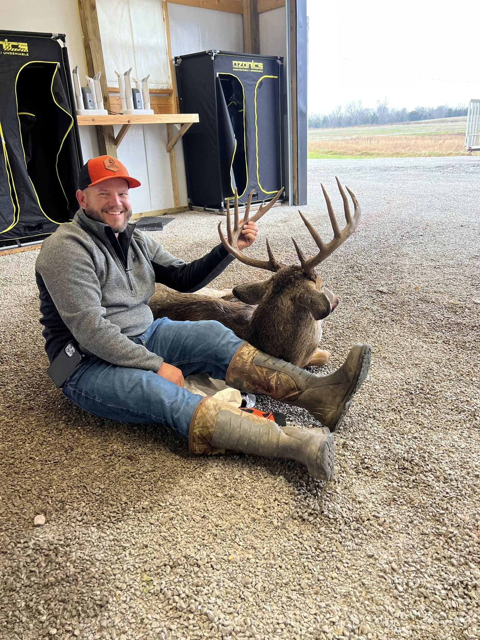 Man in orange hat and hunting gear sits with a large buck.
