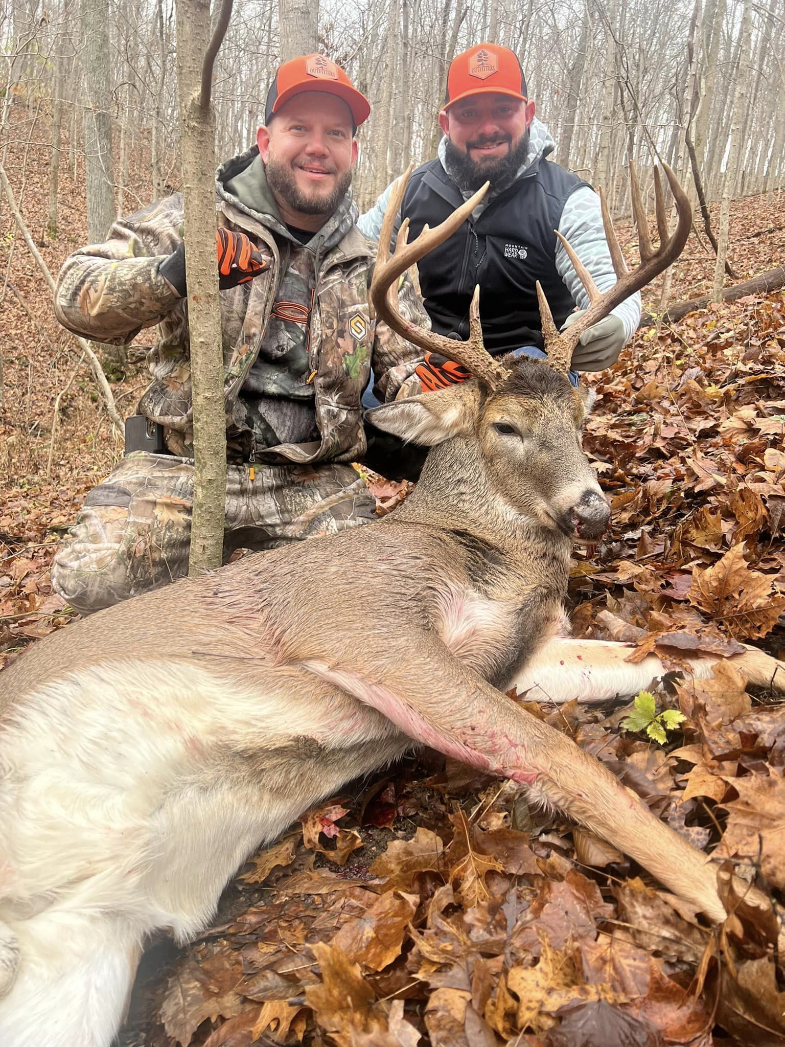 Two hunters pose with a large buck on a wooded hillside.