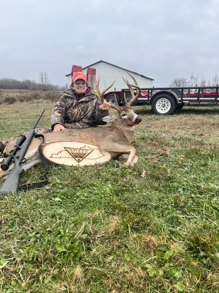 Hunter poses with a large buck next to a trailer and barn.
