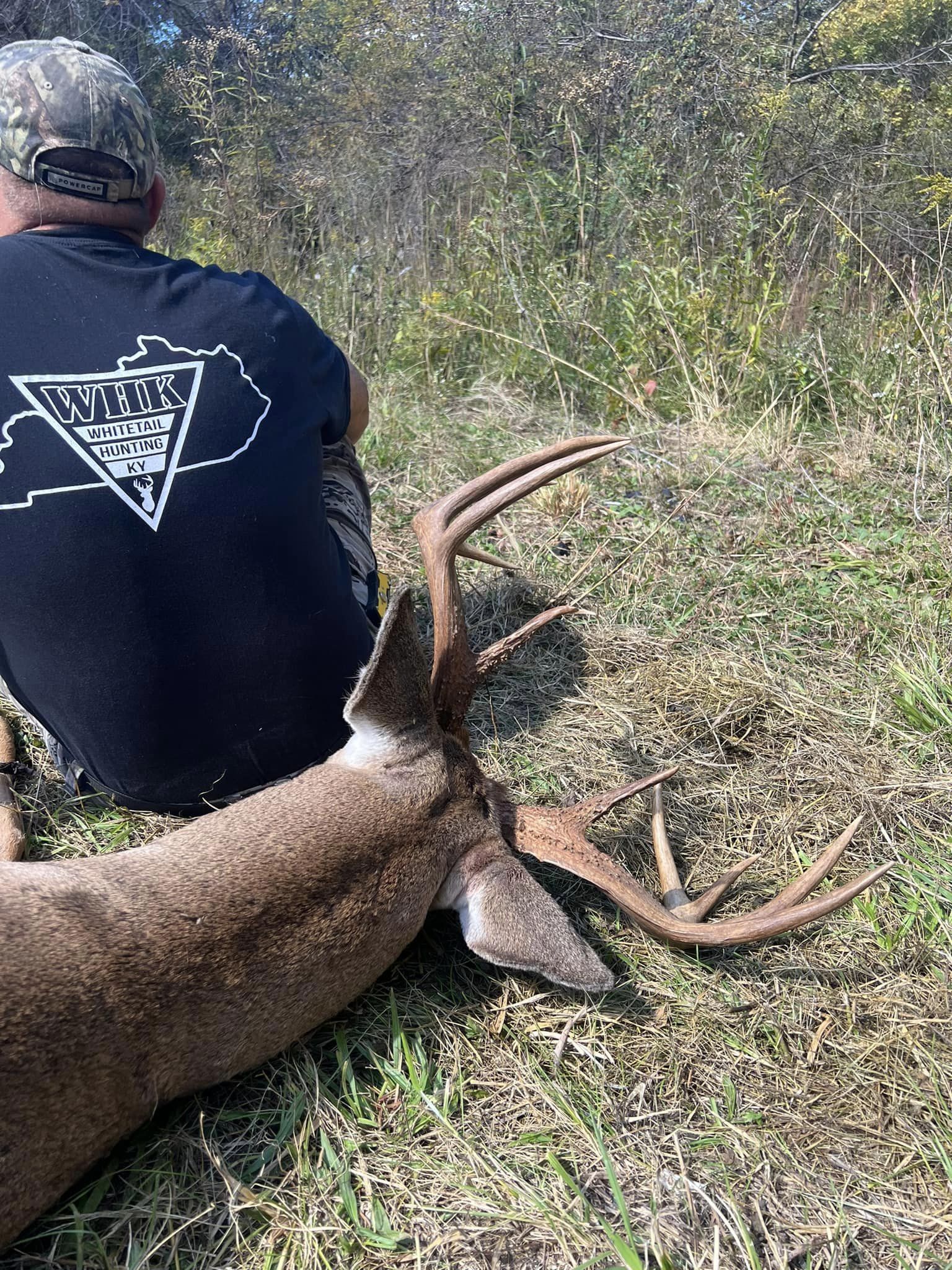 A hunter wearing a Whitetail Hunting Kentucky shirt sits by a downed buck.