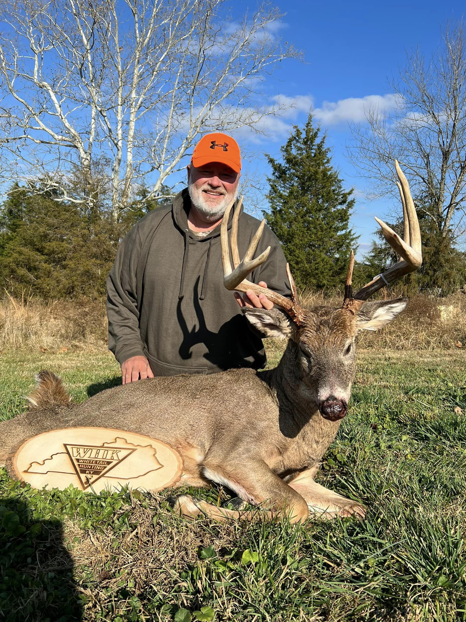 Man in orange hat poses with a large buck on a grassy field.