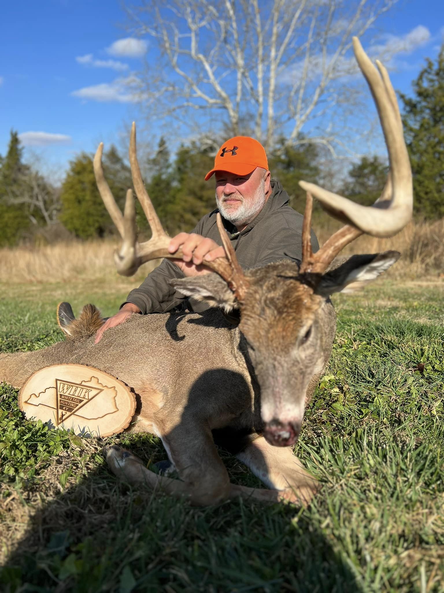 A hunter poses with a large whitetail buck beside a wooden Whitetail Hunting Kentucky logo.