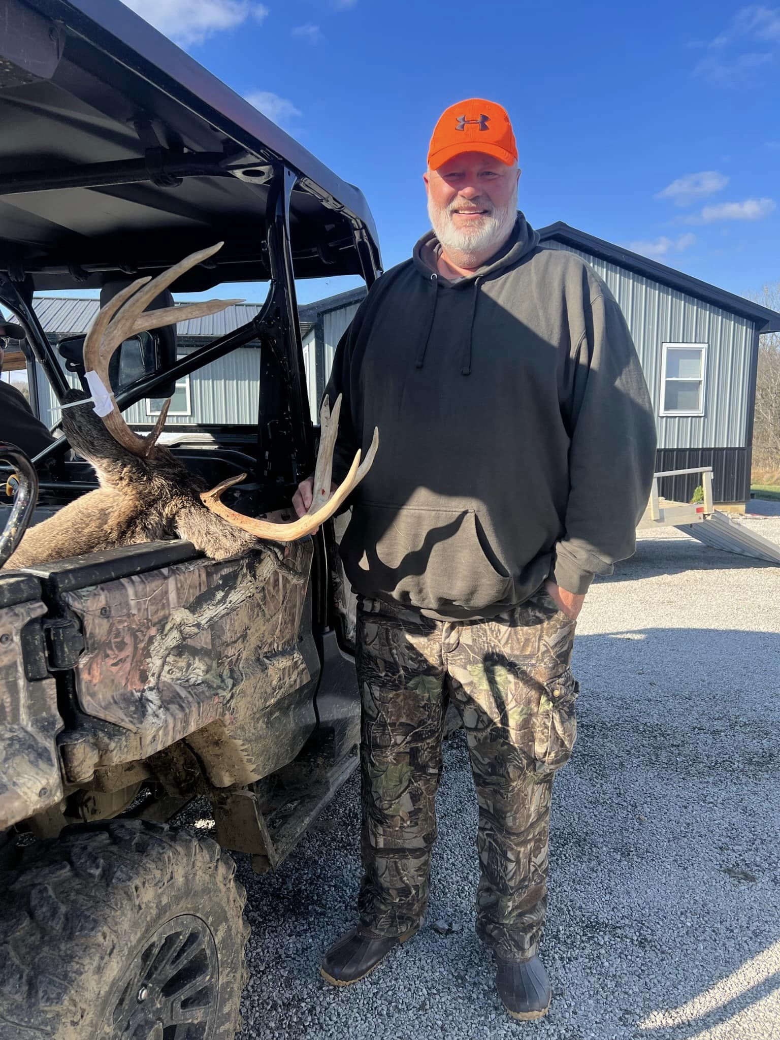 A hunter displays a harvested buck loaded onto a UTV in front of a metallic building.
