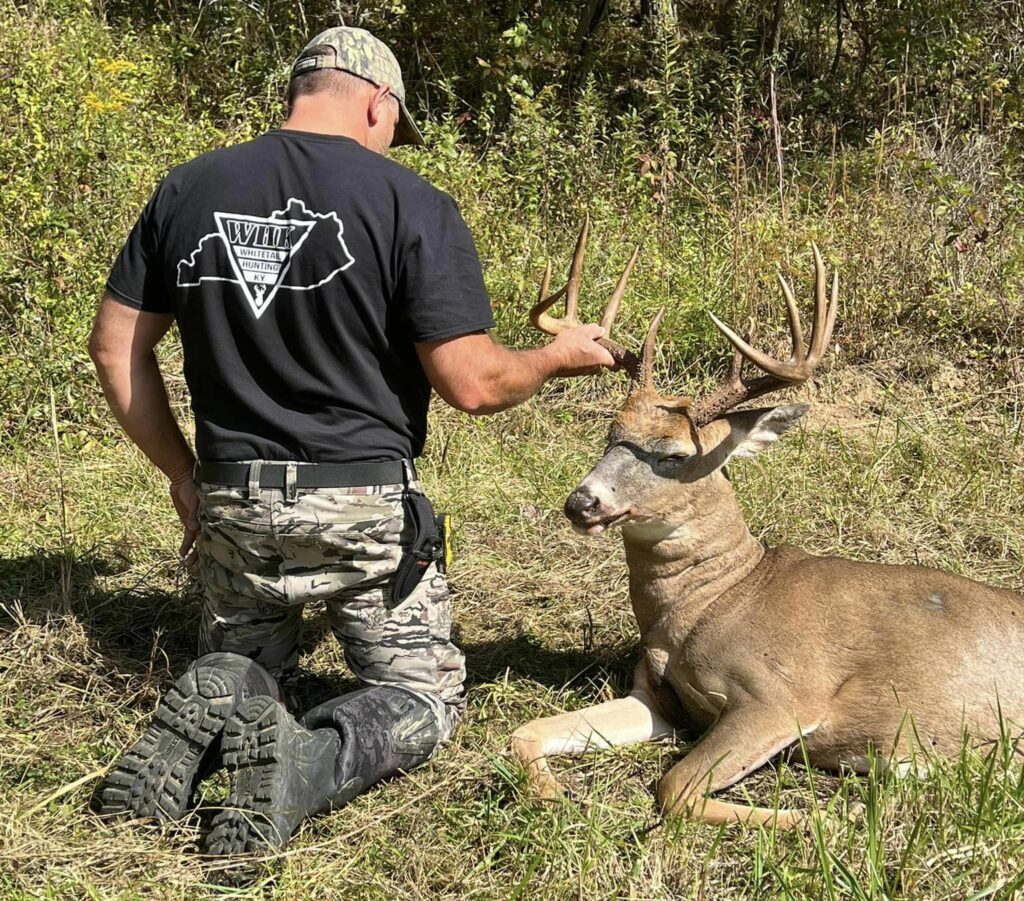 Proud hunter kneeling beside a trophy Kentucky whitetail buck in a green field.