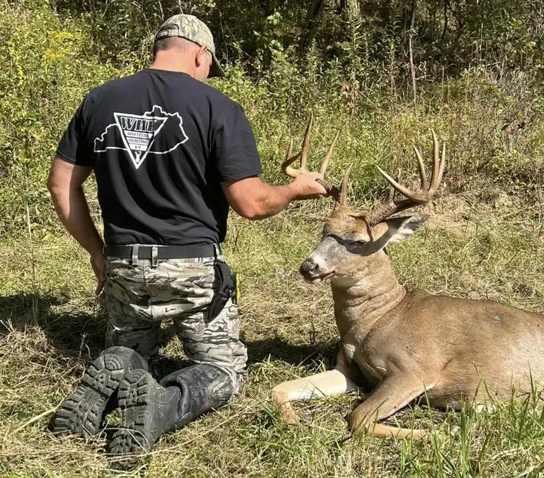 Proud hunter kneeling beside a trophy Kentucky whitetail buck in a green field.